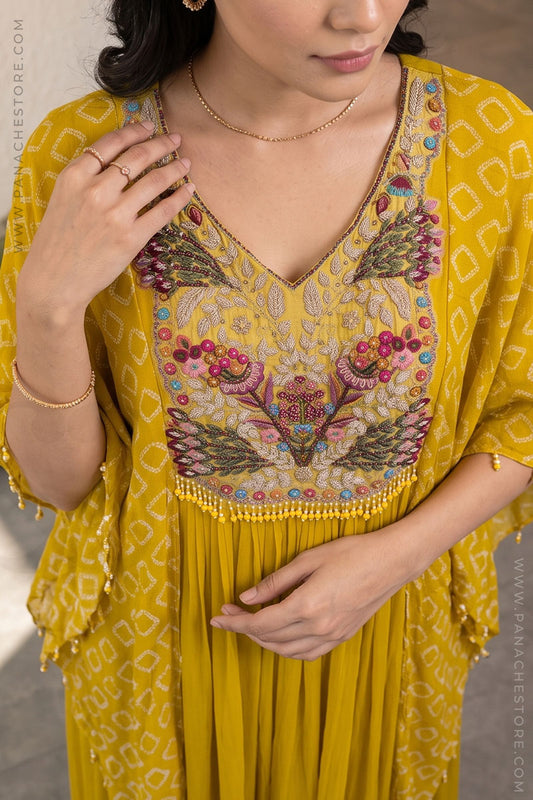 A woman wearing a mustard yellow kaftan dress with colorful floral embroidery on the bodice and a pleated skirt. The dress features a geometric pattern and tassel trim.