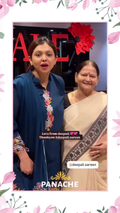 A woman in a navy blue embroidered kurta stands next to an older woman in a beige saree, both smiling in front of a dark background with a red floral decoration. The photo is framed with pink floral borders and includes social media text and the brand name PANACHE.