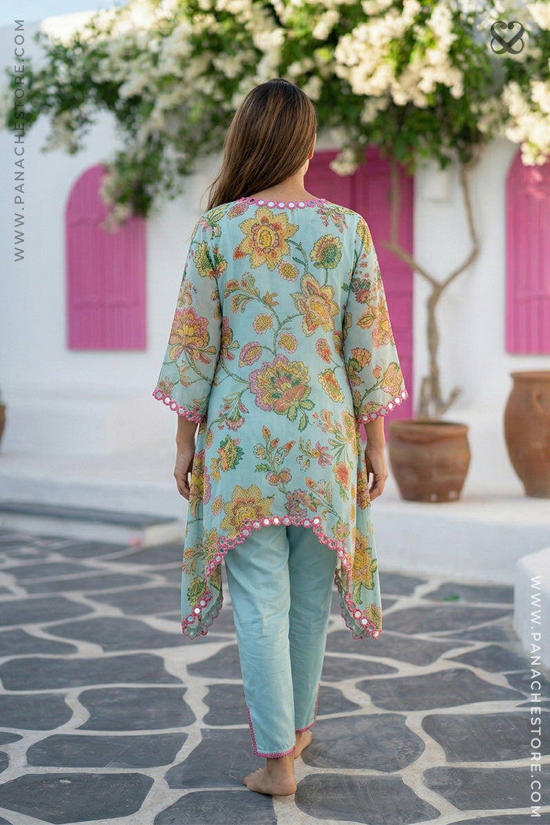 Woman wearing a floral dress and light blue pants in an outdoor setting with pink doors and white flowers.