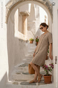 Woman in a beige dress standing in a sunlit alleyway with white walls and potted plants.