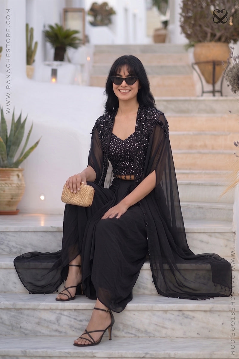 Woman in a designer black color embellished shrug cape and sequin crop top with black skirt sitting on outfoor steps