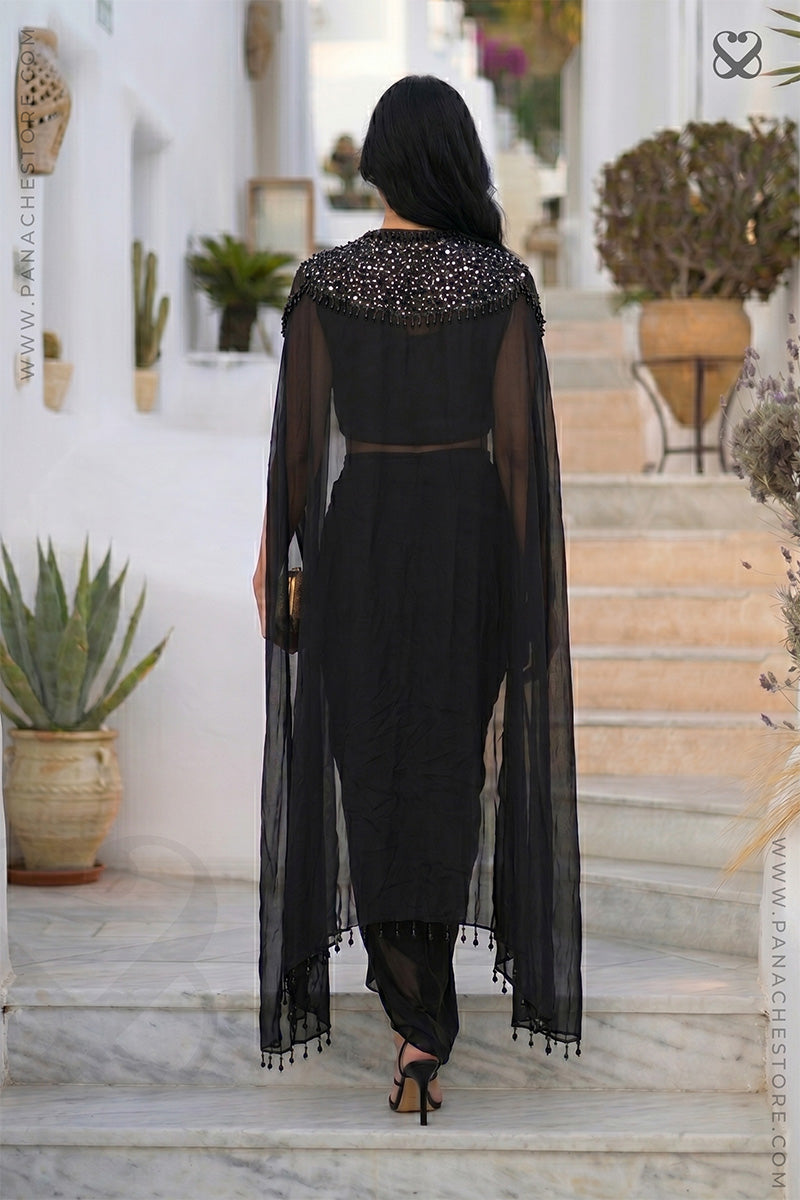 Woman in a black designer embellished cape shrug with black skirt standing in outfoor steps back pose