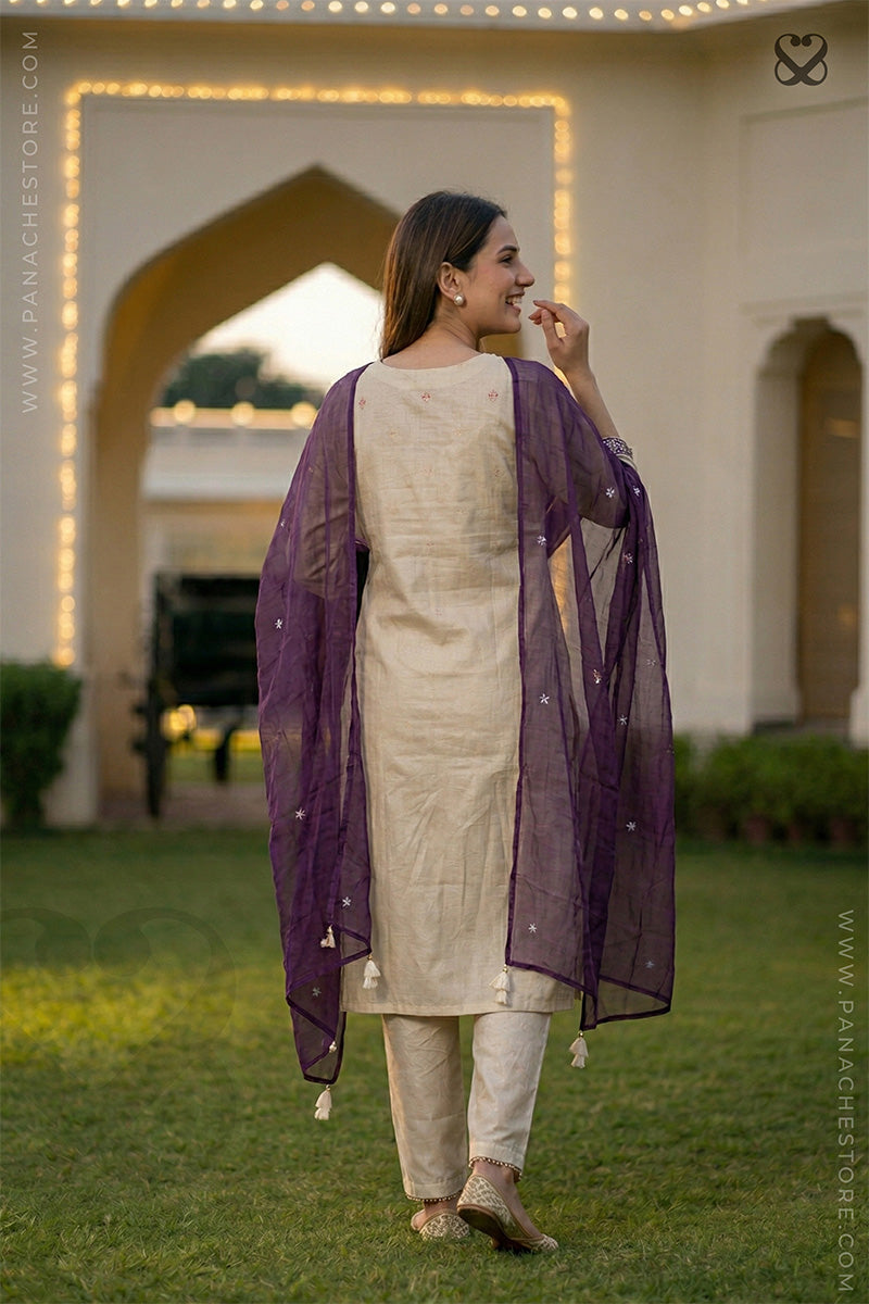 Woman in cream white slawar suit set with purple dupatta standing on grass posing back