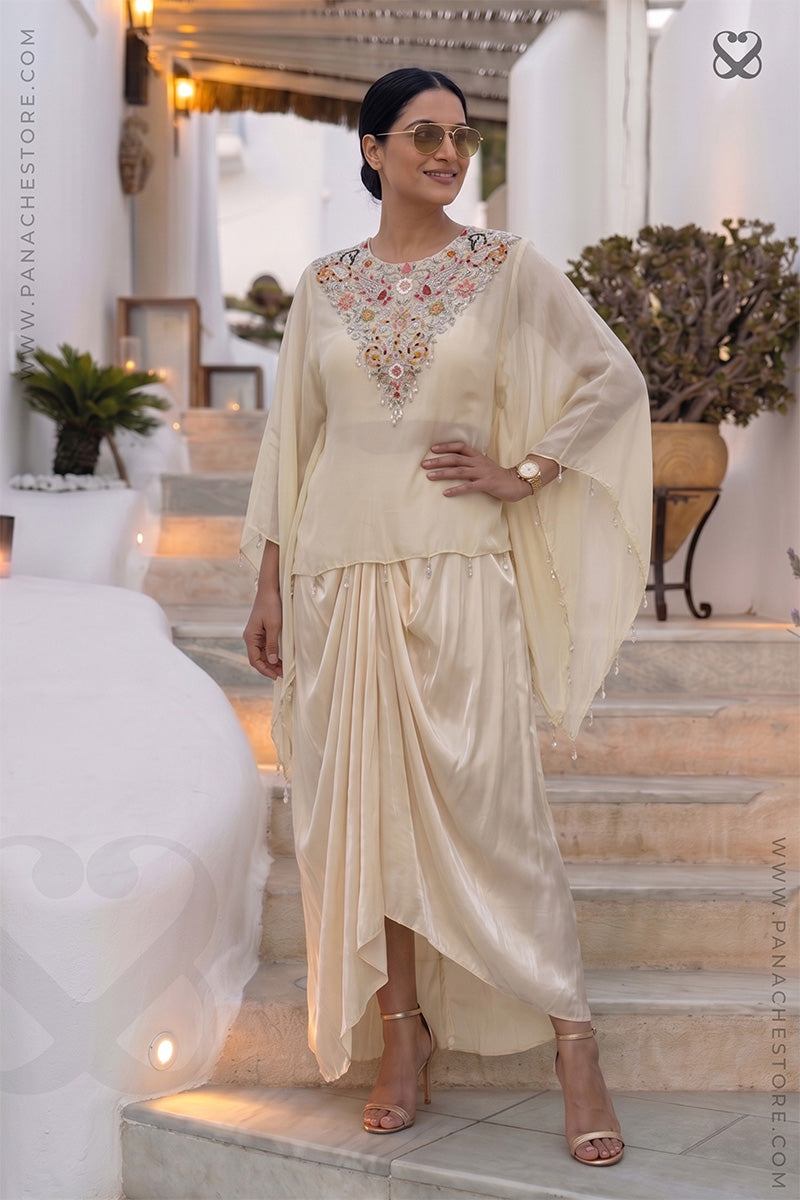 Woman in a cream color traditional kaftan with desiger embellished neckline and dhoti style skirt standing in an outdoor setting.