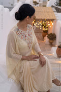 Woman in a cream color traditional kaftan with desiger embellished neckline and dhoti style skirt sitting in an outdoor setting.