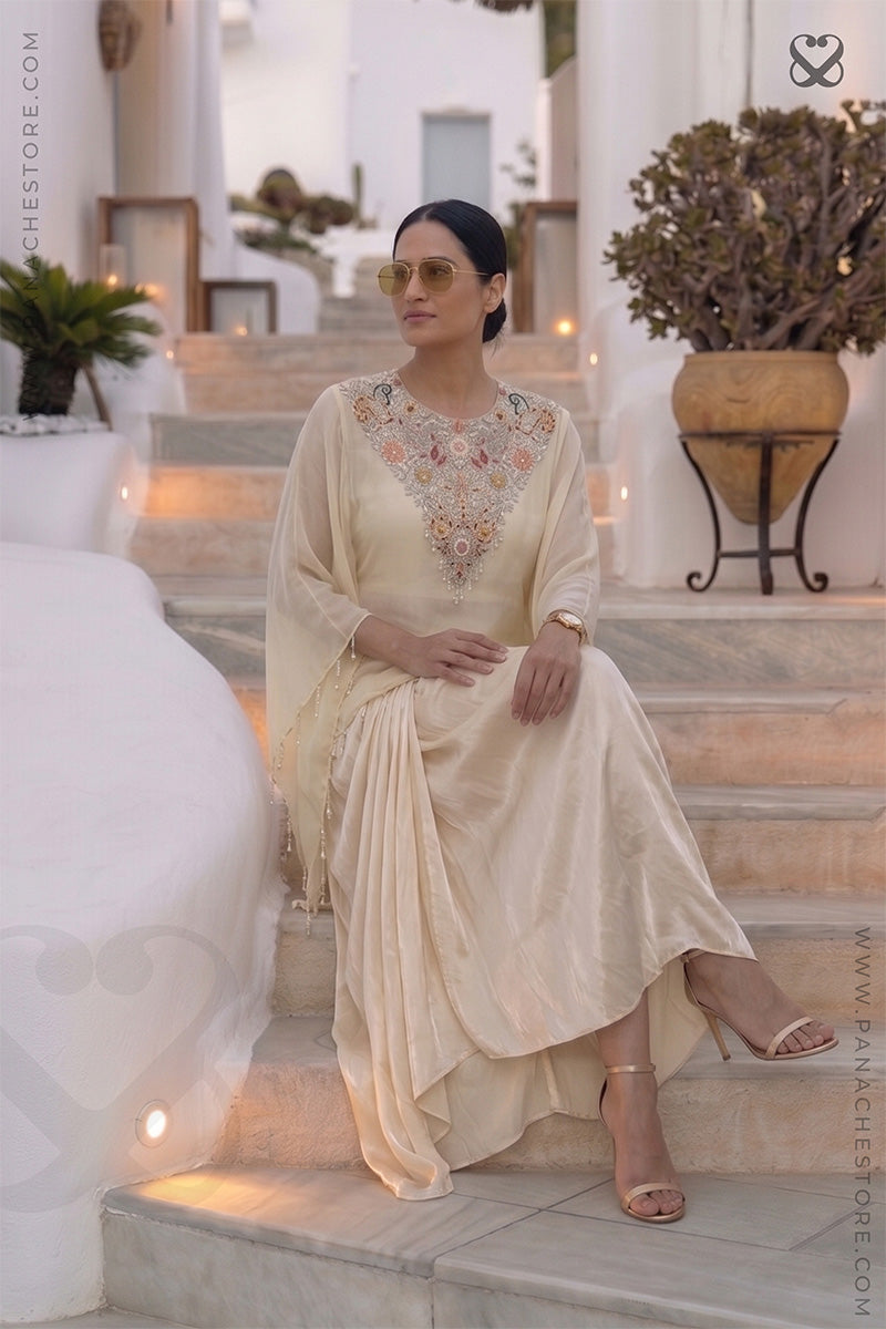 Woman in a cream color traditional kaftan with desiger embellished neckline and dhoti style skirt sitting in an outdoor setting.