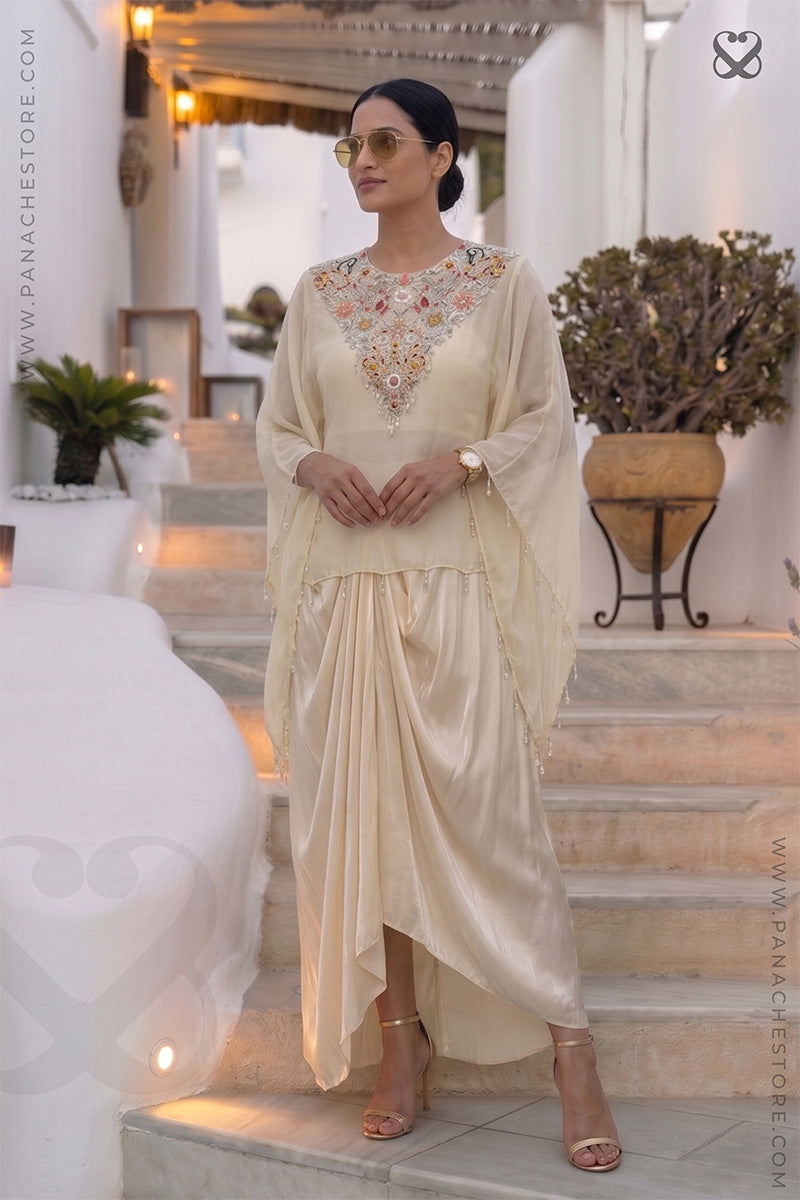 Woman in a cream color traditional kaftan with desiger embellished neckline and dhoti style skirt standing in an outdoor setting.