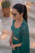 Woman in a green traditional outfit with embellishments, wearing sunglasses and gold earrings, standing outdoors.