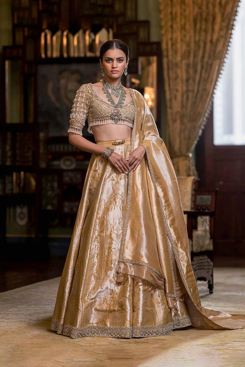 Woman in a traditional gold and beige outfit with intricate designs in a decorated room.