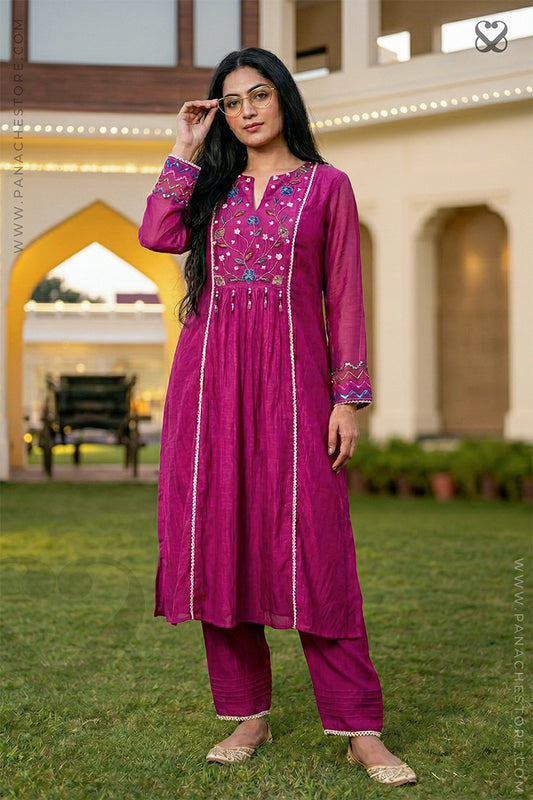 Woman in a pink traditional outfit standing outdoors with a building and greenery in the background.