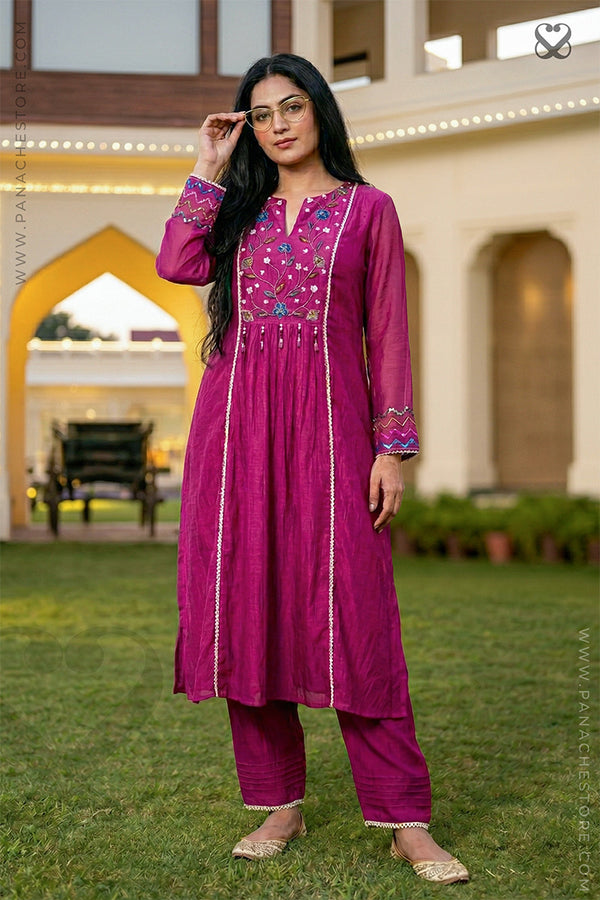 Woman in a pink traditional outfit standing outdoors with a building and greenery in the background.