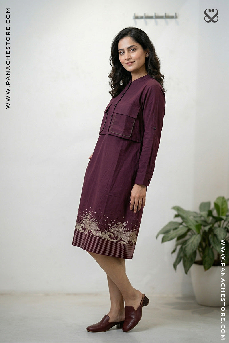 Woman wearing a maroon dress with lace details, standing against a white wall with a plant in the corner.