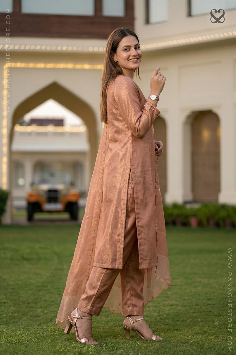 Woman in a tissue silk peach salwar suit with dupatta standing side pose in a garden