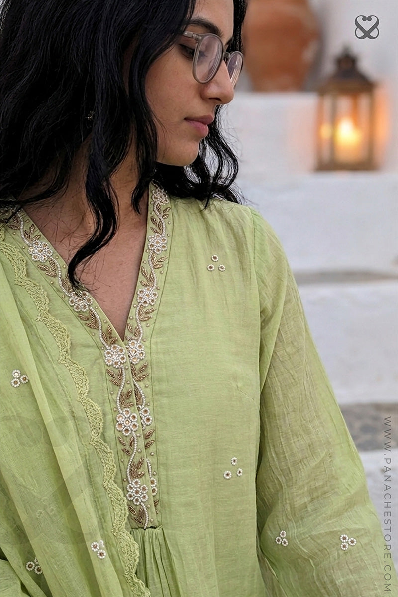 Woman wearing a light green traditional outfit with lace details, standing in a softly blurred indoor setting.