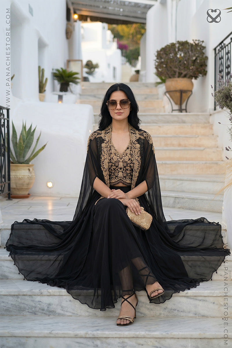 Woman in a designer cape, floral crop top and black skirt sitting on steps with plants in the background