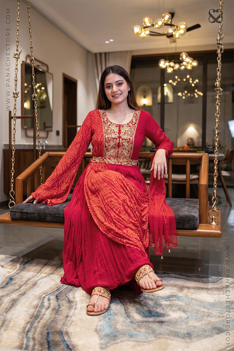 Woman in a red modern traditional dress with gold embroidery and bandhani print sitting on a swing in a decorated room.