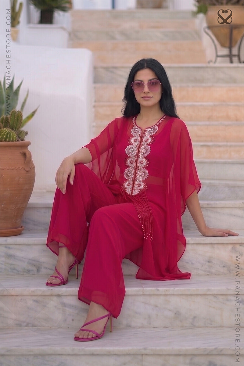 Woman in a red-pink kaftan outfit with white embroidery sitting on steps outdoors.