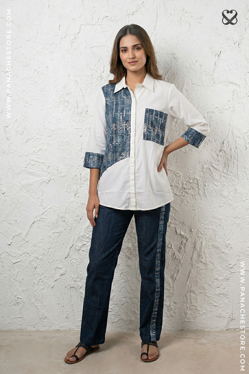 Woman wearing a white and blue patterned shirt with dark pants against a textured white wall.