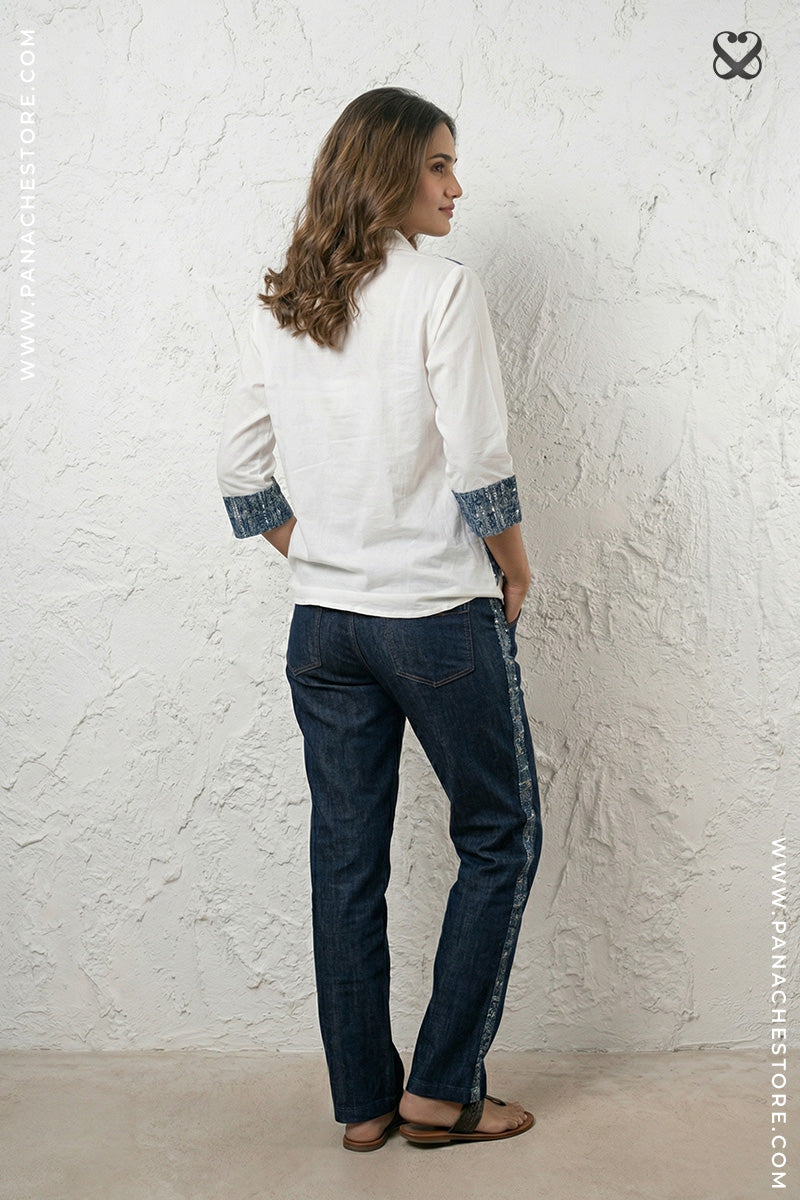 Woman wearing a white shirt with blue sleeves and dark jeans against a textured white wall.