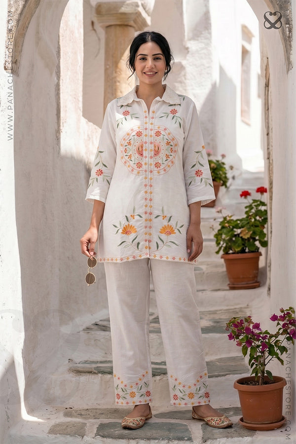 Woman wearing a white embroidered outfit standing in a sunlit outdoor setting with plants.