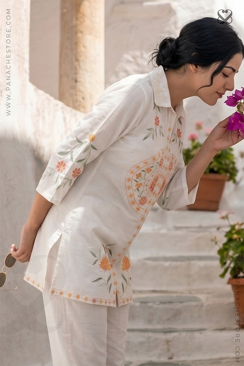 Woman in a white floral outfit holding a purple flower outdoors.