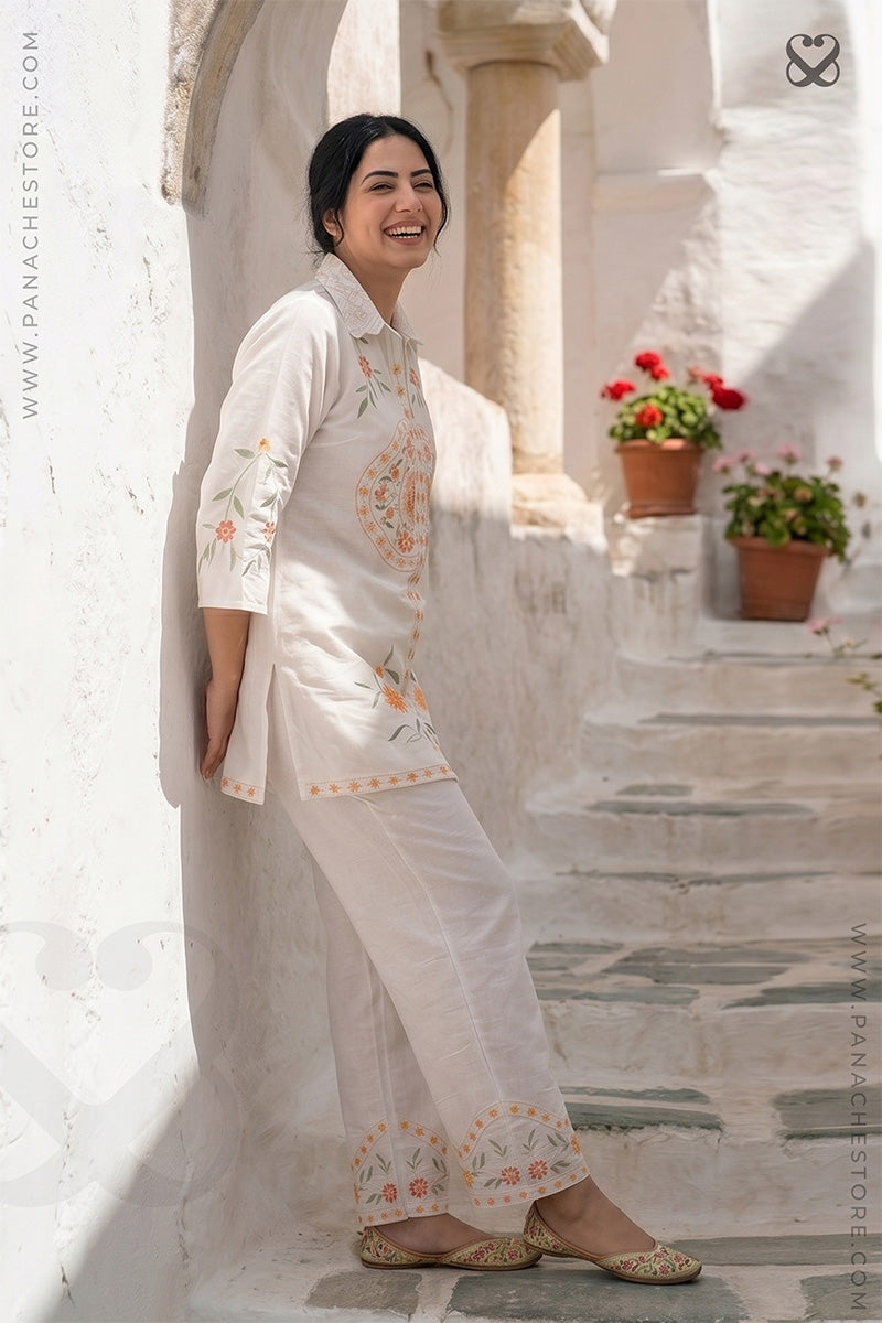 Woman in a white embroidered outfit standing on a stone staircase with flowers in the background.