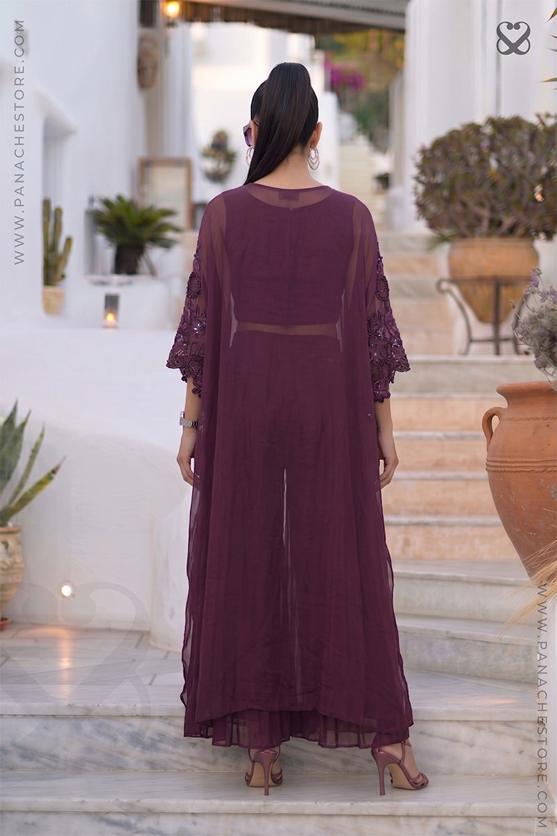 Woman in a burgundy-wine kaftan outfit standing on steps with decorative plants and architecture in the background.