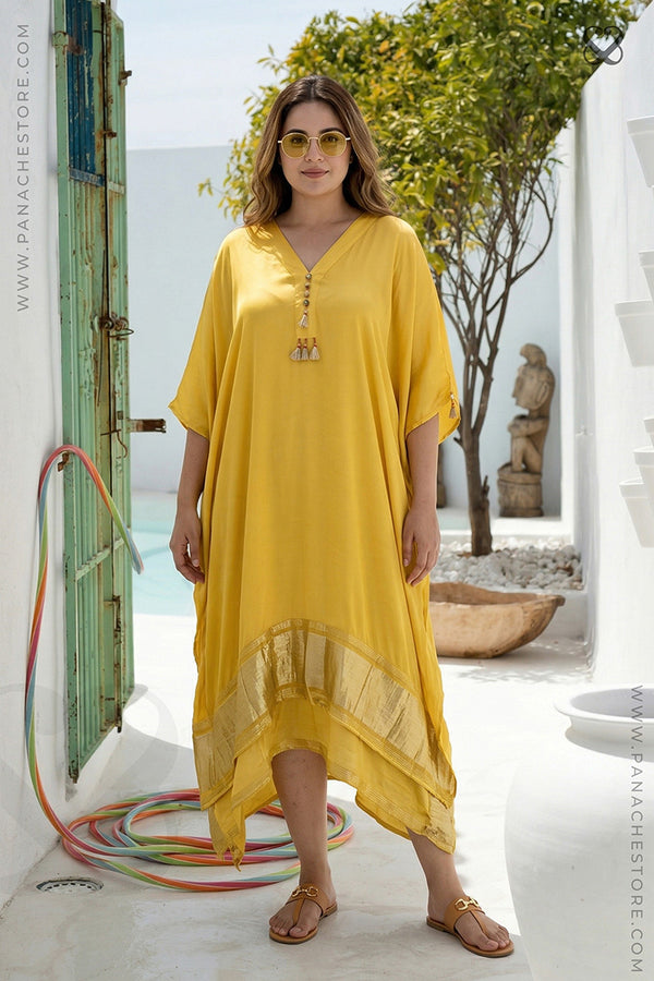 Woman wearing a yellow dress standing in a sunlit outdoor setting with greenery and architectural elements.