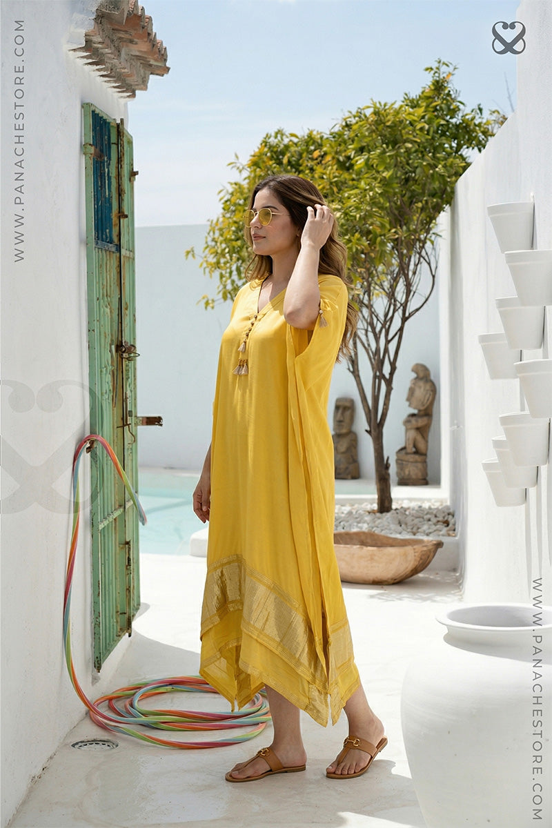 Woman in a yellow dress standing in a sunlit outdoor setting with white walls and greenery.